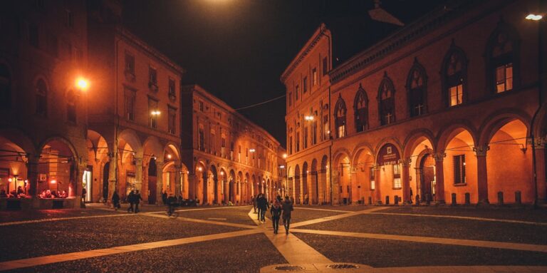 A nighttime view of a historic square in Bologna, Italy, with warm streetlights illuminating arched buildings and people walking across the cobblestone streets.