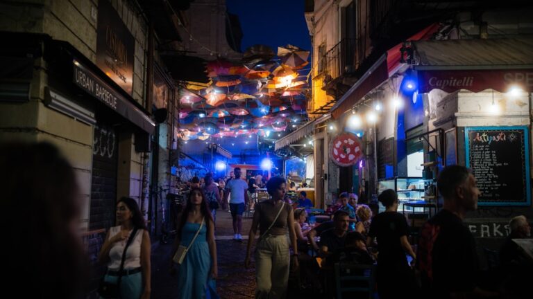 Charming night street in Europe with colorful umbrellas hanging above, cozy restaurants, and people enjoying outdoor dining. The warm lights, vibrant decorations, and lively crowd create a festive Mediterranean atmosphere perfect for summer evenings.